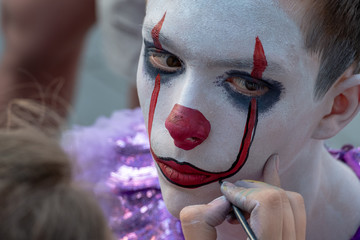 Street makeup. The image of a clown or mime. Costume for carnival or holiday halloween. Cosplay at a horror party. Artist's hand with a brush paints the face of a teenager guy. Close up.