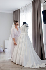Fototapeta premium Young woman in silk bathrobe touching her wedding dress and smiling while standing near the window. Wedding day. Beauty, fashion. Happiest bridal day. Enjoy every moment.