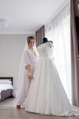 Fototapeta premium Young woman in silk bathrobe touching her wedding dress and smiling while standing near the window. Wedding day. Beauty, fashion. Happiest bridal day. Enjoy every moment.