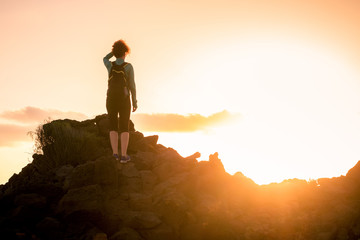 Back view of a sporty trekker girl standing on the rocks, looking at the horizon. Woman with backpack walking in the evening at sunset. Backlight silhouette. Sport adventure freedom and hiking concept