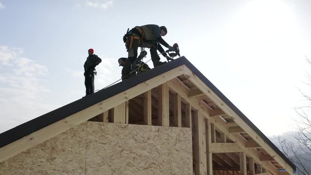 Aerial zooming in view of three builders in safety harness installing soft tiles on the roof using hydraulic hammer. Residential house under construction