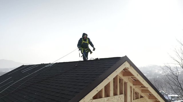 Aerial zooming in view of builder in safety harness installing soft tiles on the roof. Wooden frame house under construction. Bright sunny day