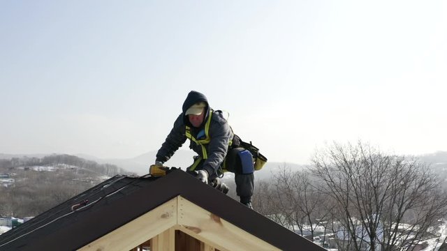 Aerial portrait of professional builder in safety harness installing soft tiles on the roof. Wooden frame house under construction. Bright sunny day