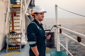 Marine Deck Officer or Chief mate on deck of offshore vessel or ship , wearing PPE personal protective equipment - helmet, coverall. He holds VHF walkie-talkie radio in hands.