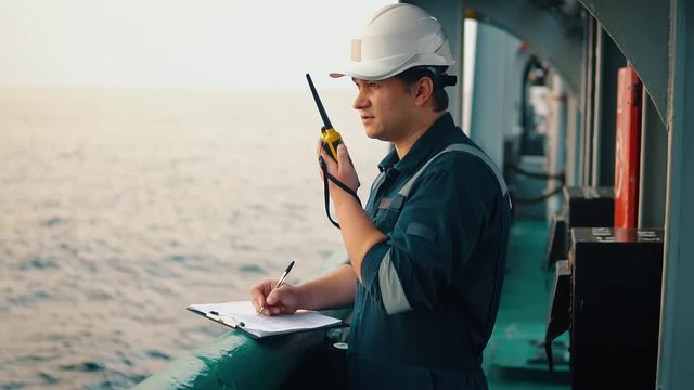 Marine chief officer or chief mate on deck of ship or vessel. He fills up ahts vessel checklist. Ship routine paperwork. He holds VHF walkie-talkie radio in hands.