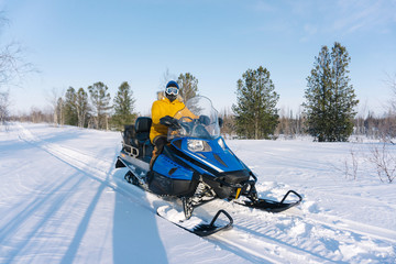 young guy in a yellow warm jacket sits on his snowmobile and rides through an empty snowy meadow surrounded by trees on a frosty winter day