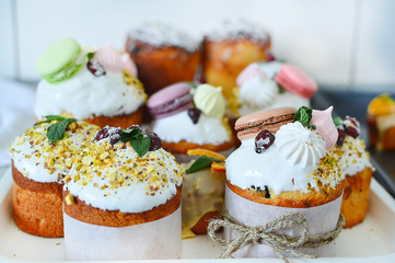 Easter cakes and multi-colored eggs decorated with macaroons, mint, wild rose with white icing, lilac branches on a white wooden background. Easter.