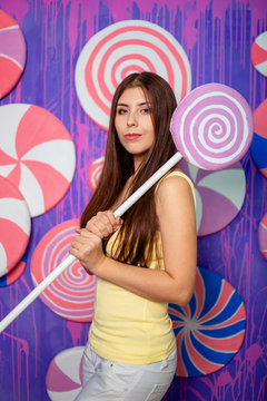 A beautiful, young girl with a big toy candy in her hands, stands on an abstract violet-blue background of sweets and lollipops.