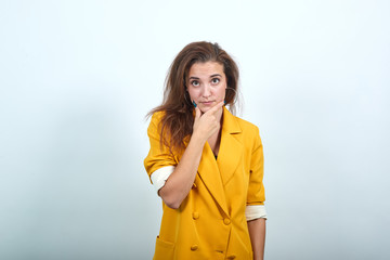 Attractive young woman in yellow jacket keeping hand on chin, looking directly isolated on gray...