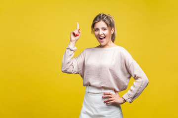 Portrait of inspired beautiful young woman with fair hair in casual beige blouse standing with finger up, rejoicing at solution found, creative idea. indoor studio shot isolated on yellow background