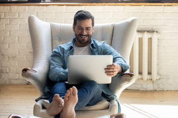Smiling relaxed young man sit on armchair using laptop laughing