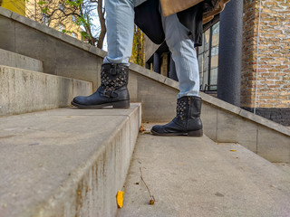 Woman going forward one step at a time climbing step staircase with black punk boots on a concrete steps. Autumn fall, blue jeans and yellow jacket