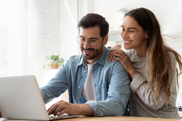 Smiling millennial couple using laptop looking at screen at home