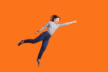 Fototapeta na wymiar Superhero. Portrait of joyous pretty woman with brown hair in long sleeve shirt jumping high with one stretched arm, feeling to be superman flying up. indoor studio shot isolated on orange background