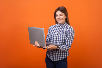 Portrait of cheerful woman with brown hair in checkered casual shirt and denim standing holding laptop computer, looking at camera with toothy smile. indoor studio shot isolated on orange background