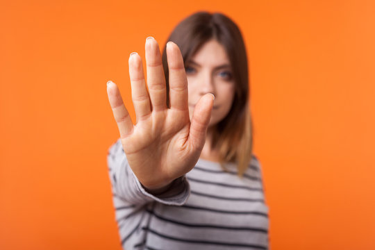 Portrait of young brunette woman in long sleeve striped shirt standing with serious look and showing stop hand gesture at camera, refusal or prohibition. indoor shot isolated on orange background