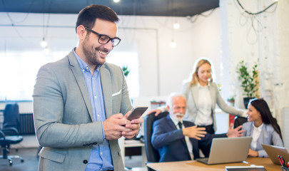 Smiling businessman using phone in the office with colleagues in the background.