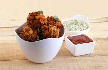 Pakoda, or pakora, a traditional, popular, and vegetarian Indian snack and side dish, with onion as the main ingredient, in a basket, and coconut chutney and tomato ketchup, in a bowl on a wooden back