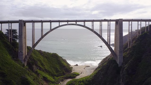 Fantastic aerial footage of one of the most beautiful arch bridges standing between two green hills in front of the Pcific ocean. Drone moves slowly under the bridge span. Cars drive the bridge. 4K