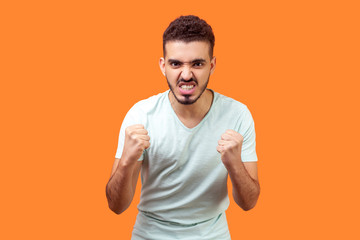 Portrait of furious irritated brunette man with beard in white t-shirt standing with raised fists and clenched teeth, experiencing strong aggression, anger management problems. indoor studio shot