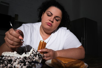 Depressed overweight woman eating sweets in kitchen at night