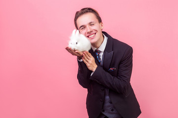 Portrait of excited man in elegant suit and with stylish hairdo hugging adorable white rabbit bunny...