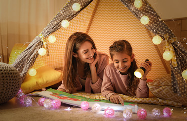 Mother and daughter with flashlight reading book in play tent