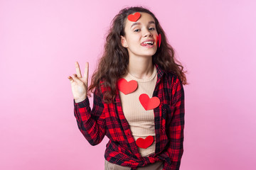 Valentines Day. Portrait of playful cute teen girl with curly brunette hair and paper heart stickers on her face and clothes showing tongue and gesturing victory, peace. studio shot, pink background
