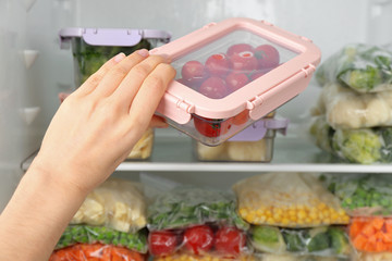Woman putting container with tomatoes in refrigerator with frozen vegetables, closeup