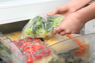 Woman putting plastic bag with broccoli in refrigerator with frozen vegetables, closeup