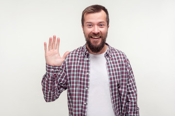 Fototapeta premium Hello! Portrait of good-natured positive bearded man in casual plaid shirt waving hand and friendly smiling at camera, making hi welcome gesture. indoor studio shot isolated on white background
