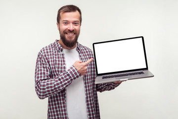 Portrait of positive bearded man in casual plaid shirt holding laptop and pointing at empty screen, smiling satisfied with computer app. indoor studio shot isolated on white background, place for ad - obrazy, fototapety, plakaty