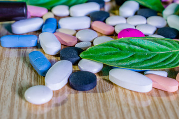lots of tablet and capsules in different colors kept on a wooden table with green herbal leaves    