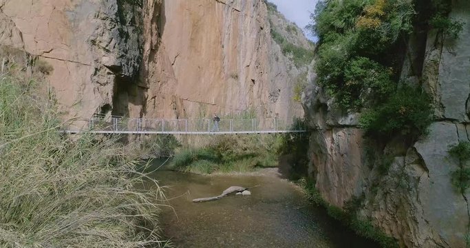 Dron avanzando y ascendiendo dentro de r&iacute;o entre paredes de piedra vertical y persona mayor caminado en puente colgante 