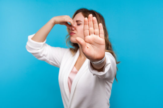 Bad smell. Portrait of woman with wavy hair in white jacket pinching her nose and showing stop gesture, expressing disgust to unpleasant odor, fart gases, her eyes closed with abhor. blue background