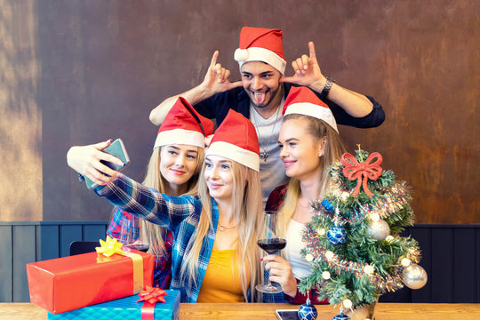 Happy young friends wearing Santa claus hat enjoying Christmas party taking selfie with funny faces at home sharing it on social media