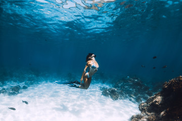 Fototapeta premium Freediver girl with fins glides over sandy bottom with fishes in blue ocean