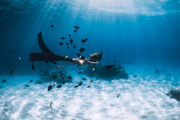 Freediver girl with fins glides over sandy bottom with fishes in blue ocean