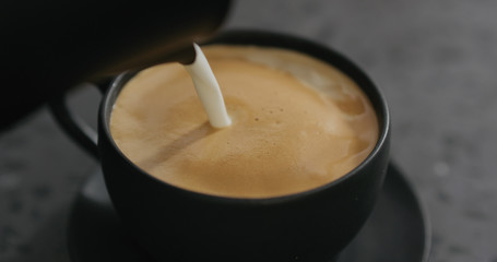 closeup pouring steamed milk into cappuccino in black cup on terrazzo countertop
