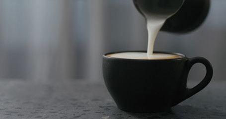 pouring steamed milk into cappuccino in black cup on terrazzo countertop