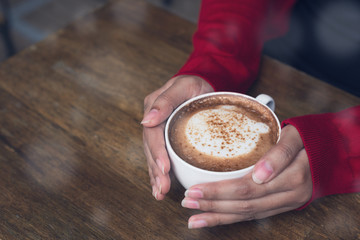 Woman's hands in red cadigan holding cup of cappucino coffee in winter.