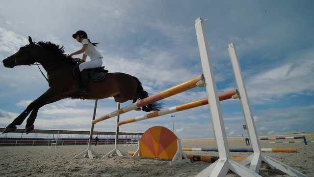 Horsegirl riding strong brown horse jumping fence in outdoors sandy parkour riding arena. Competitive rider training jumping over obstacles at the arena. Equestrian sport. Slow motion, low angle view