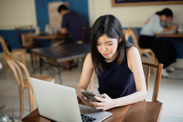 Asian girl freelance using smartphone while working with her laptop in coffeeshop.