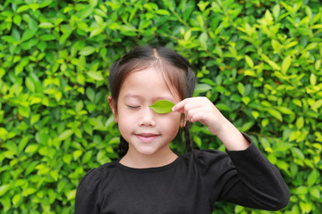 Portrait of happy little Asian child girl holding a green leaf closing left eye in green garden background.