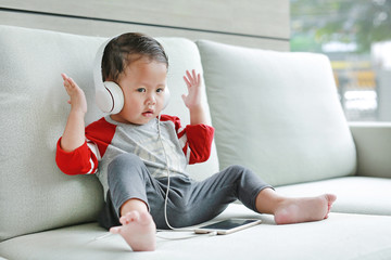 Adorable little baby boy sitting on sofa and listening music at headphones by smartphone. Kid using earphones to relaxing indoors.