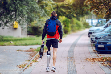 Rear view of sporty caucasian handicapped man with artificial leg, in sportswear and backpack walking on the street. In hands is artificial leg.