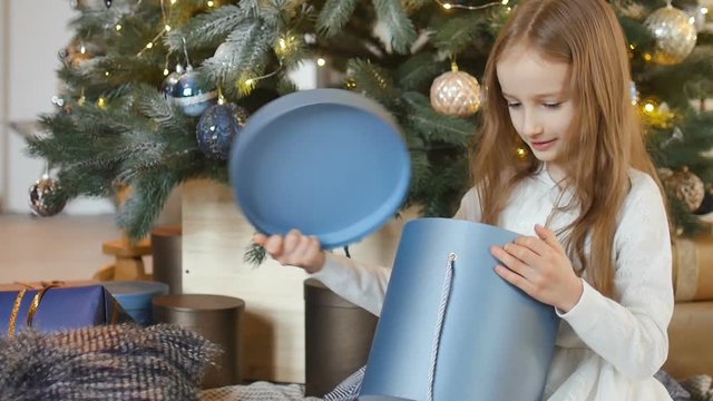 Cute blond girl is opening dark blue gift box she recieved sitting near christmas tree, happy childhood and xmas celebration concepts