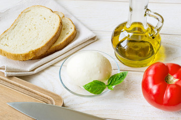 Italian mozzarella cheese, red tomato and olive oil in glass jar near sliced bread on a white rustic wooden table. Healhy eating and vegetarian food.