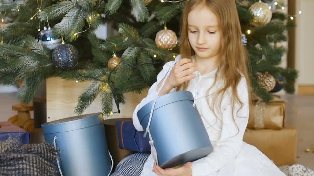 Cute blond girl is opening dark blue gift box she recieved sitting near christmas tree, happy childhood and xmas celebration concepts