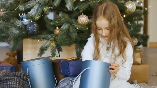 Cute blond girl is opening dark blue gift box she recieved sitting near christmas tree, happy childhood and xmas celebration concepts